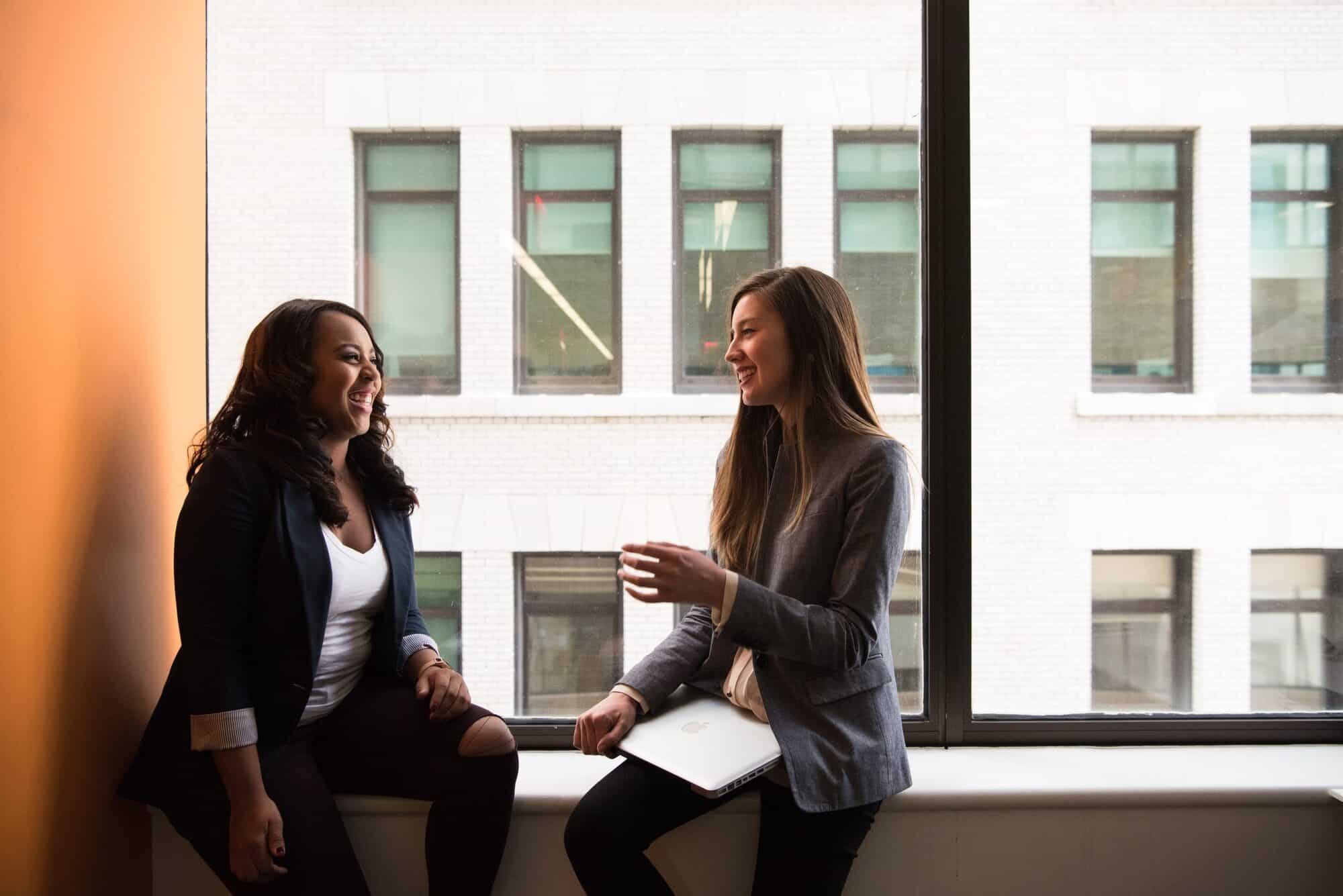 Two women in business attire sitting on a window ledge having a discussion.