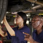 A female apprentice working underneath a car under instruction.