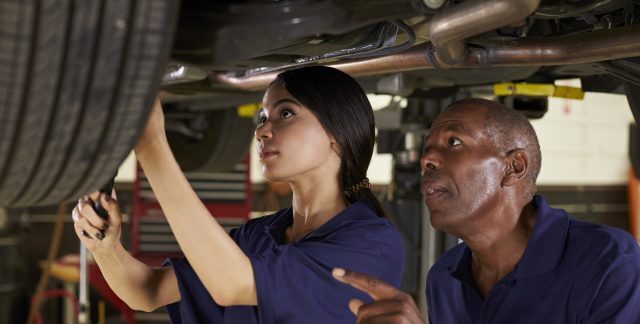 A female apprentice working underneath a car under instruction.