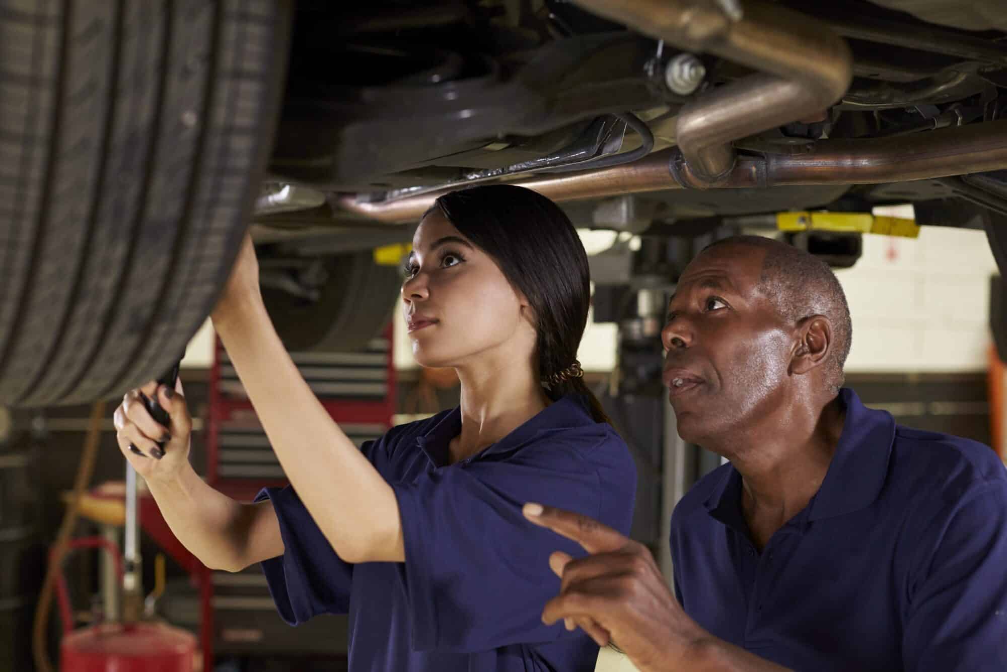 A female apprentice working underneath a car under instruction.