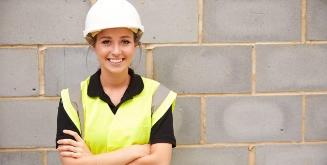 A young woman in a hard hat and high visibility jacket by a concrete brick wall