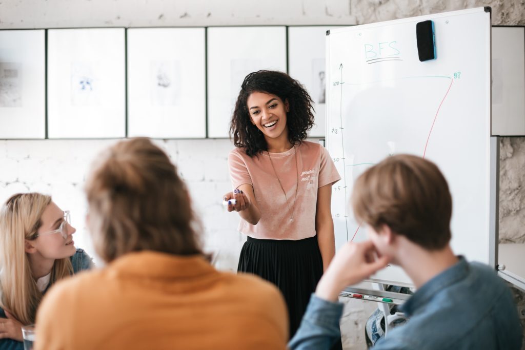 A woman in front of a whiteboard talking to a class.