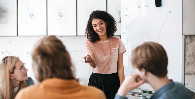 A woman in front of a whiteboard talking to a class.