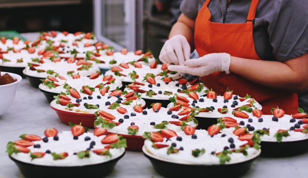 A person in a red apron and gloves prepares a number of identical desserts decorated with strawberries