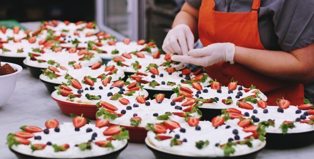 A person in a red apron and gloves prepares a number of identical desserts decorated with strawberries
