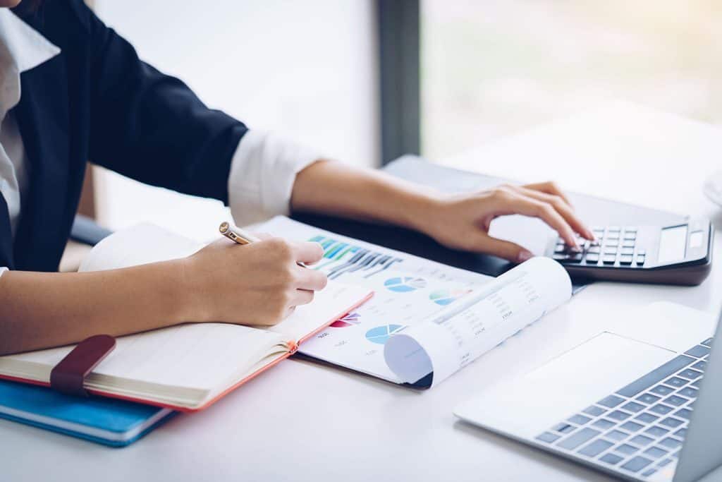 A person writes in a notepad while looking at a laptop and charts