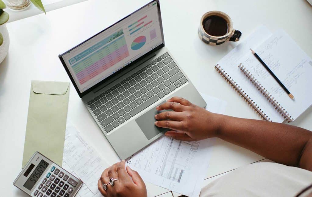 A person using a laptop. On the screen are reports. On the desk a cup of coffee