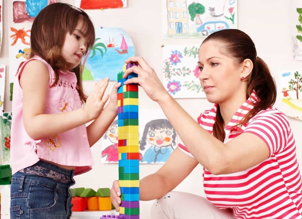 A childcare practitioner helps a child build a duplo lego tower. Behind them are children drawings and paintings