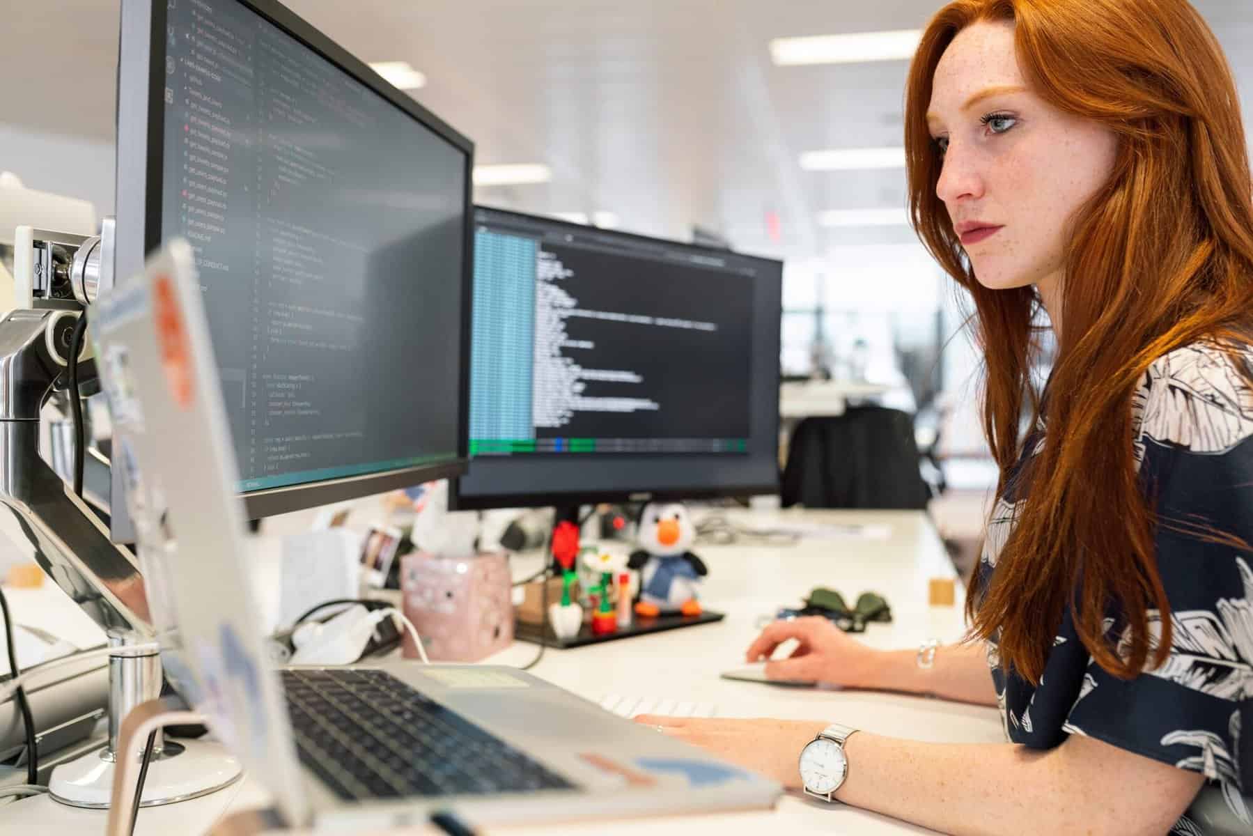 A young woman codes while looking at two monitors and a laptop