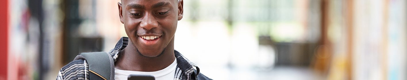 A smiling student looking at his phone while walking down a corridor holding some folders