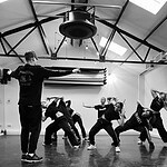 A group of dancers in casual attire practicing in a studio with high ceilings and exposed beams. The choreographer stands at the front, directing the dancers who are mid-movement, striking dynamic and expressive poses. The lighting casts dramatic shadows on the floor.