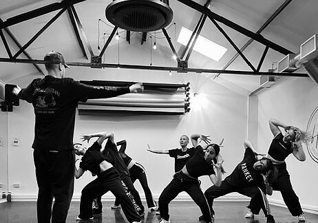 A group of dancers in casual attire practicing in a studio with high ceilings and exposed beams. The choreographer stands at the front, directing the dancers who are mid-movement, striking dynamic and expressive poses. The lighting casts dramatic shadows on the floor.