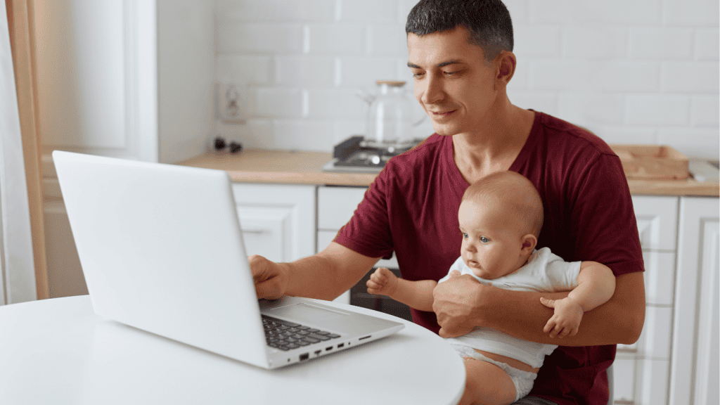A man is seated at a white table, working on a laptop with a baby in his arm. The baby is wearing a white outfit, and both are in a bright kitchen with white cabinets and a clean, minimalistic aesthetic.