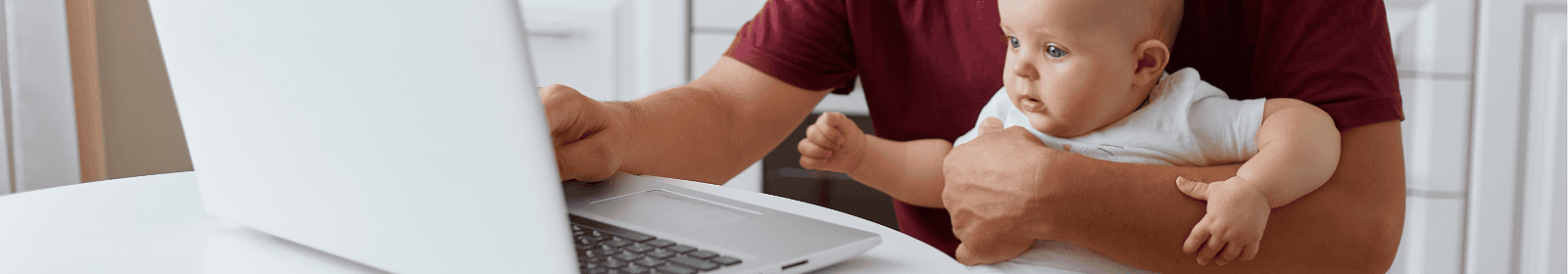 A man is seated at a white table, working on a laptop with a baby in his arm. The baby is wearing a white outfit, and both are in a bright kitchen with white cabinets and a clean, minimalistic aesthetic.