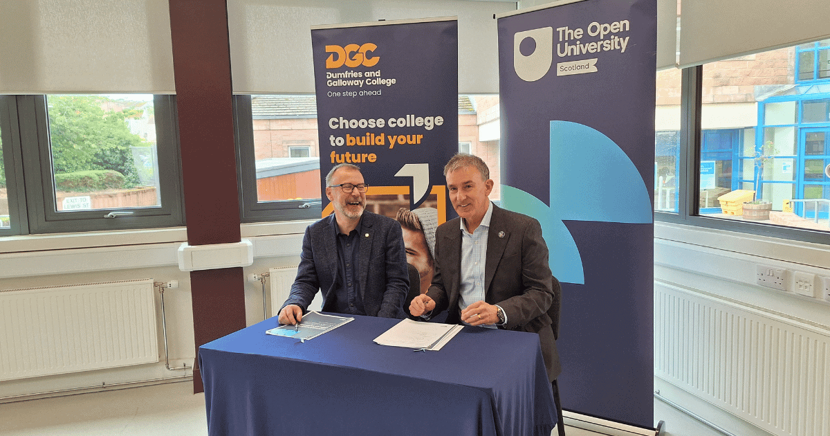 An eye-level shot shows two men sitting at a table with a blue tablecloth, signing documents in front of two banners. On the left, a man wearing a dark blue patterned blazer, a dark blue button-up shirt, and glasses smiles and looks to his left. He holds a pen in his right hand above a document on the table. Behind him is a banner with the Dumfries and Galloway College logo and the text "Choose college to build your future." On the right, a man in a dark brown blazer and light blue button-up shirt looks at the camera, holding a pen above a document on the table. Behind him is a blue banner with The Open University Scotland logo. Windows in the background show a building and trees outside.